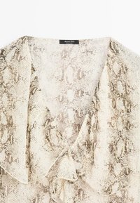 Pytontryckt blus med ett mjukt, genomskinligt tyg, V-ringning och flödande, volangdetaljer fram. Färgpaletten inkluderar beige och brunt.