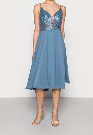 Blauwe midi-jurk met een lijfje van pailletten en diepe V-hals, gecombineerd met een vloeiende chiffon rok, gedragen met zilverkleurige sandalen met hoge hak.