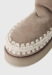 Pantofii din suede bej cu cusături decorative bej, accente țesute de-a lungul marginii și o talpă din cauciuc. Prim-plan al zonei de vârf.