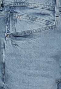 Tela de denim azul claro con una textura suave, que presenta un pequeño bolsillo trasero y un detalle de botón plateado, costura a lo largo del borde.