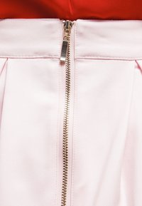 Gonna rosa con cerniera frontale con dettagli dorati, pieghe in vita e tessuto dalla texture liscia. Sopra si intravede parzialmente una maglietta rossa.