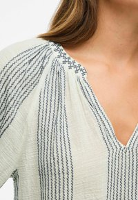 Femme portant un haut crème avec des rayures verticales cousues en marine et un décolleté en V avec des détails de col brodés, gros plan sur l'épaule et la poitrine.