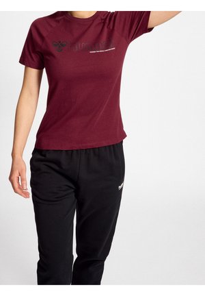 Person trägt ein burgunderfarbenes Kurzarmshirt mit "hummel"-Logo und schwarze Hose, steht vor einem einfarbigen weißen Hintergrund.