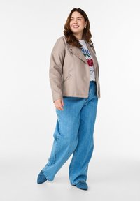 Taupefarbene Bikerjacke aus Kunstleder mit asymmetrischem Reißverschluss, kombiniert mit blauen, übergroßen Jeans und spitzen blauen Schuhen.