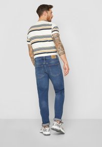 Slim-fit τζιν παντελόνι σε σκούρο μπλε χρώμα με ξεθωριασμένη υφή, συνδυασμένο με λευκό πουκάμισο κοντομάνικο ριγέ που φέρει πορτοκαλί, μαύρες και γκρι ρίγες.