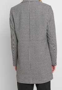 Manteau long à motif pied-de-poule noir et blanc, coupe ajustée, fente unique au dos et col cranté. Texture lisse.