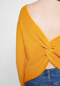 Gele gebreide trui met een open rug en gedraaide knoopdetail. Heeft een brede halslijn en een gestructureerd steekpatroon. Gecombineerd met spijkerbroek.