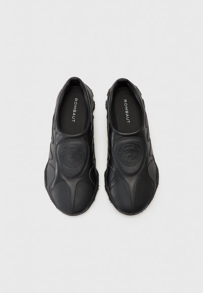 Zapatos negros tipo slip-on hechos de cuero suave, con un logo redondo en relieve en la parte superior y una suela de goma texturizada. Diseño plano.