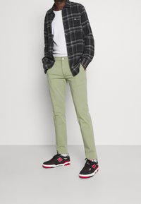 Pantalones verdes, camiseta blanca, camisa de cuadros negra y gris, zapatillas negras con detalles en rojo. De pie con las manos en los bolsillos sobre un fondo neutro.