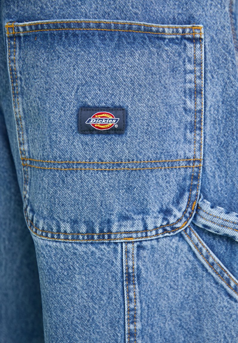 Blaue Denim-Overalls mit großer Brusttasche, Doppelnaht und einem Dickies-Logo-Patch. Der Stoff hat ein strukturiertes, abgenutztes Aussehen.