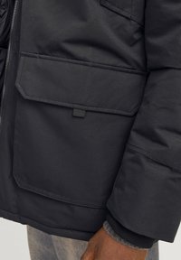 Veste matelassée noire avec une surface texturée, une grande poche frontale et des poignets côtelés. Le matériau semble durable et résistant aux intempéries.