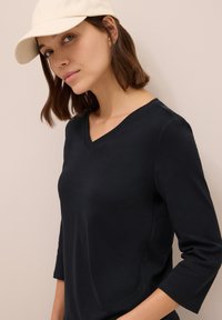 Haut noir à manches longues et col en V, fabriqué dans un tissu lisse, associé à une casquette de couleur claire. Le visage du modèle est partiellement visible, sur un fond neutre.
