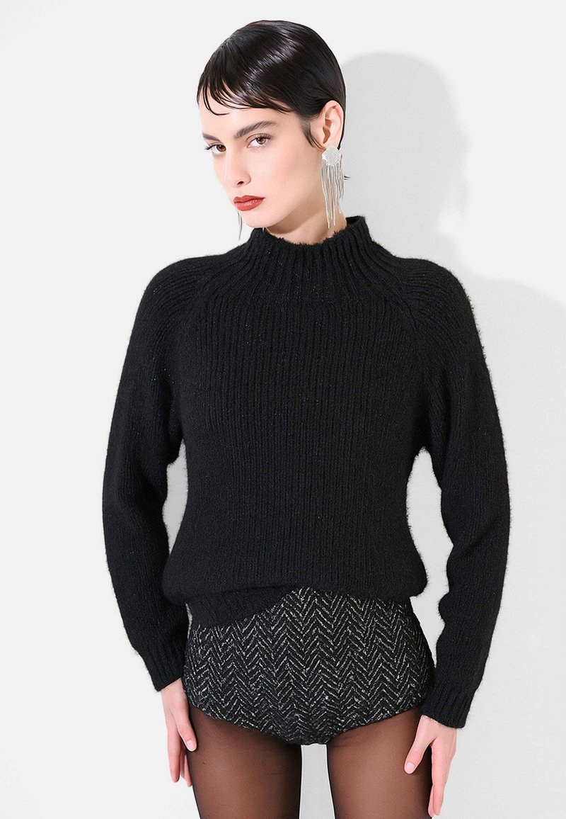 Maglione nero lavorato a maglia con collo alto e texture a coste, abbinato a pantaloncini scuri con fantasia. Il modello indossa orecchini lunghi argentati.