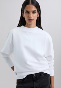 Woman with long dark hair wears a white sweatshirt and black jeans, posing with one hand partially in pocket against a plain background.