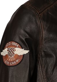 Ärmel einer braunen Lederjacke mit einem runden Air Crew-Abzeichen, das Flügel, ein Schild und den Text "JCC-1984 Airforce LXXIII 05" zeigt.