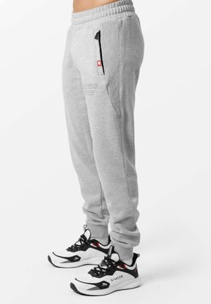 Lichtgrijze joggingbroek met elastische tailleband en zijzak met rits gedragen met witte en zwarte sneakers bij een staand persoon.