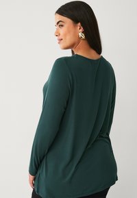 Long-sleeve, dark green shirt made of smooth fabric with a relaxed fit and a rounded neckline, featuring a subtle drape at the back.