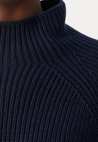 Gros plan sur un pull à col roulé en maille côtelée bleu marine foncé sur le cou et l'épaule d'une personne.