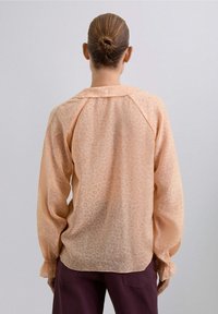 Mujer con el cabello recogido en un moño, vistiendo una blusa de manga larga en melocotón claro con un sutil estampado de leopardo y pantalones morados oscuros, de espaldas.