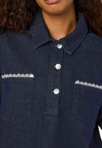 Temno modra jeans srajca z ovratnikom, srebrnimi gumbi in čipkastimi poudarki ob žepih. Teksturiran material s klasičnim dizajnom.