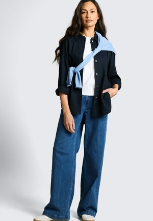 Femme aux longs cheveux foncés portant une chemise blanche, une chemise noire à boutons, un jean bleu à jambes larges et un pull bleu clair noué autour des épaules.