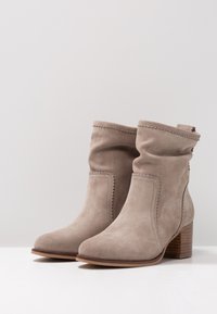 Bottines en daim taupe avec une tige incurvée, des détails festonnés et un talon en bois superposé. Texture lisse et design à bout arrondi.