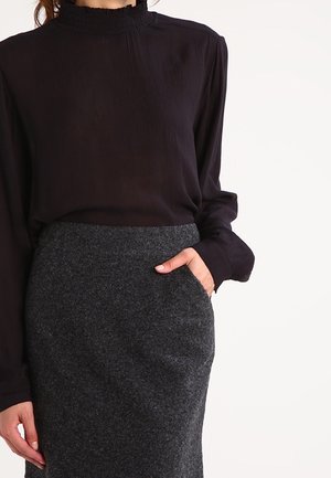 Femme portant un chemisier noir à manches longues avec un col à volants et une jupe texturée gris foncé taille haute avec des poches.