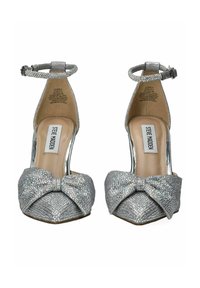 Zapatos de tacón alto plateados con una textura brillante, que presentan un detalle de lazo en la punta y una correa en el tobillo. Tacón transparente y plantilla acolchada.