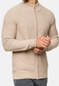 Hombre vestido con un suéter de punto beige con cremallera, con las manos entrelazadas al frente, combinado con pantalones gris oscuro.