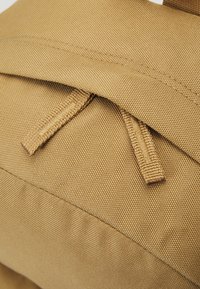 Material in tela color ocra con finitura testurizzata, dotato di due linguette intrecciate e cuciture per una maggiore durata.