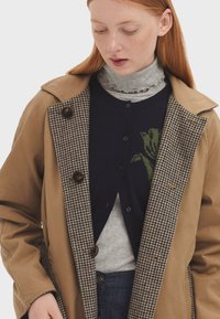 Abrigo beige con forro de pata de gallo, botones marrones, usado sobre un cárdigan azul marino con un diseño floral verde, superpuesto a un cuello de tortuga gris claro.