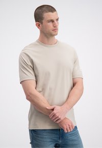 Jasnobeżowa koszulka z krótkim rękawem o okrągłym dekolcie, gładkiej fakturze i małą metką na lewym rękawie. Noszona z niebieskimi dżinsami.