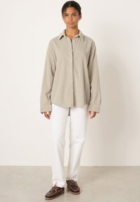 Mladá žena stojí, oblečená v béžové manšestrové košili na knoflíky, bílých kalhotách a tmavě hnědých šněrovacích botách na jednobarevném světlém pozadí.