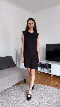 Γυναίκα με μακριά σκουρόχρωμα μαλλιά που περπατάει μέσα στο σπίτι με μαύρο αμάνικο φόρεμα και μαύρα παπούτσια Mary Jane πάνω σε στρογγυλό γκρι χαλί.