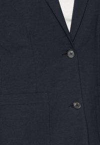 Tmavě námořnický blazer se strukturovaným designem, s klopovým límcem, jediným černým zapínáním na knoflík a dvěma předními kapsami. Hladká textura.