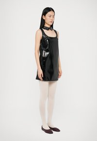 Γυναίκα με μακριά μαύρα μαλλιά, φορώντας μια χωρίς μανίκια κομψή μαύρη μίνι φόρεμα, λευκές καλσόν, καφετιά παπούτσια τύπου φλάτ και μαύρο κολάρο, που στέκεται μπροστά σε λευκό φόντο.