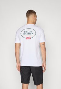 Weißes T-Shirt mit einem runden Grafikdesign, das "TRAVIS MATHEW" und florale Akzente zeigt, kombiniert mit schwarzen Shorts.