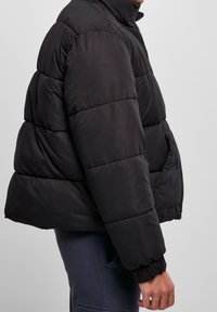 Chaqueta de plumas negra con un diseño acolchado, cuello alto, cierre de cremallera y puños elásticos, que ofrece una apariencia estructurada y aislante.
