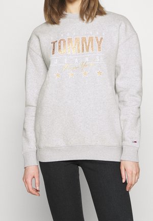 Persoon draagt een lichtgrijze sweatshirt met metalen tekst "TOMMY JEANS New York" en sterren, gecombineerd met een zwarte broek.
