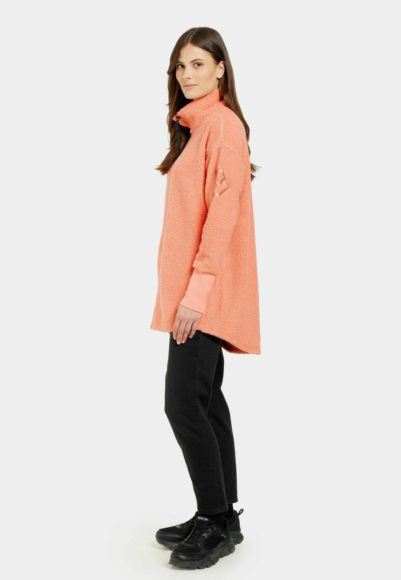 Fleece-Pullover in Koralle mit hohem Kragen und seitlichen Schlitzen, kombiniert mit schwarzen Jeans und klobigen schwarzen Schuhen. Strukturierter Stoff und lässige Passform.