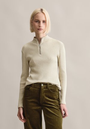 Beige geribde trui met een halve ritssluiting, gecombineerd met olijfgroene corduroy broek met voorzakken en een knoopsluiting.