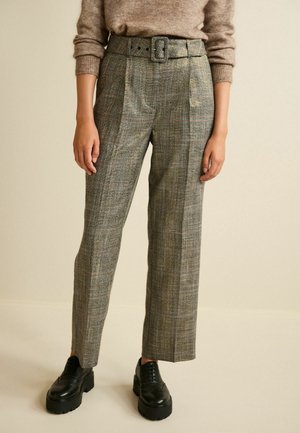 Personne portant un pantalon à carreaux taille haute avec ceinture, un pull marron et des chaussures montantes en cuir noir épaisses, debout devant un fond beige uni.