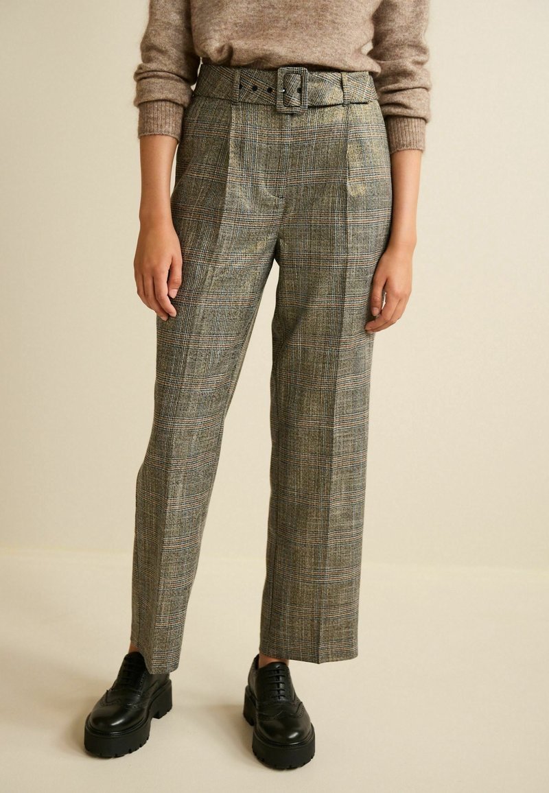 Personne portant un pantalon à carreaux taille haute avec ceinture, un pull marron et des chaussures montantes en cuir noir épaisses, debout devant un fond beige uni.