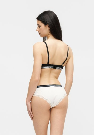 Femme aux longs cheveux noirs portant un soutien-gorge noir à bretelles et un sous-vêtement blanc bordé de dentelle, debout de dos à la caméra.