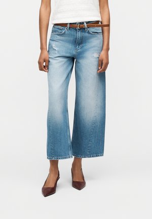 Femme portant un jean large court bleu clair avec des patches usés, une ceinture marron, un haut blanc et des talons pointus marron.