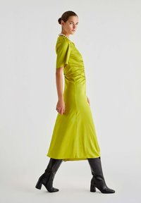 Abito in satin verde con maniche corte, corpetto aderente, fianchi arricciati, gonna a balzana lunga fino alla caviglia, abbinato a stivali neri con tacco.