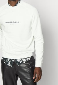Felpa bianca con il testo "BOGLIOLI", colletto rotondo, polsini a coste, abbinata a una camicia a righe con colletto e pantaloni in pelle nera.