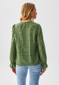 Groene kanten blouse met bloemenpatroon, lange pofmouwen en gerimpelde manchetten. Sluiting met sleutelgat aan de achterkant. Draagt lichtblauwe spijkerbroek.