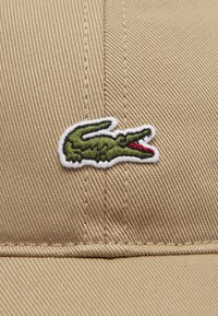 Boné bege com tecido texturizado e um logotipo de crocodilo bordado em branco, apresentando detalhes em verde e vermelho. Costura detalhada visível.
