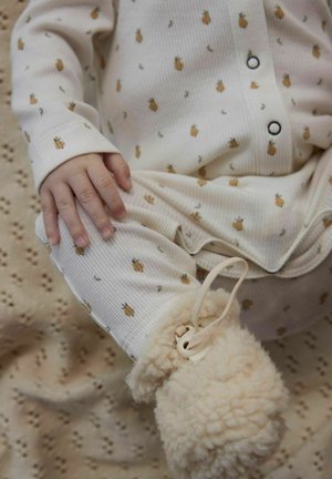 Ensemble pour bébé en tissu blanc côtelé avec de petits motifs de ananas marron. Une main repose sur le pantalon assorti, accompagné de pantoufles crémeuses et duveteuses.